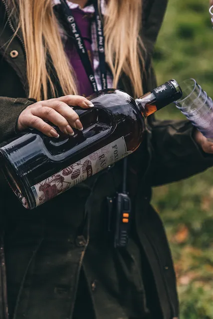 A lady pouring a glass of foxdenton gin at a game shoot featuring a dog and field lanyard in the background
